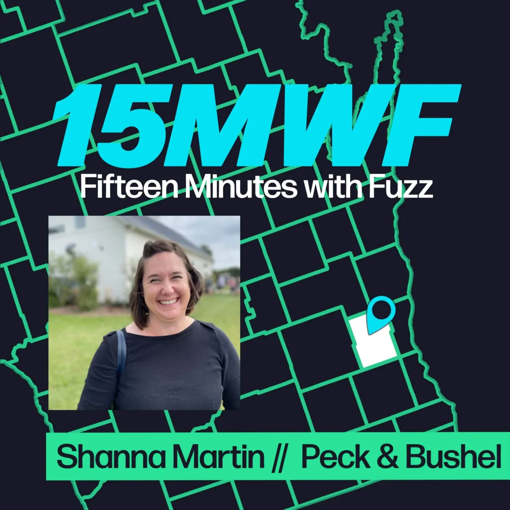 A modern design featuring the text “15MWF” in bold, bright turquoise letters at the top. Below it, the full name “Fifteen Minutes with Fuzz” is written in white. The background includes a map outline in green, with a highlighted area pinpointed in white and blue, indicating a specific location. At the bottom, there is a rectangular photo of a woman with short brown hair, smiling, and wearing a black top. Below her photo, the text “Shanna Martin // Peck & Bushel” is displayed in green on a black strip.