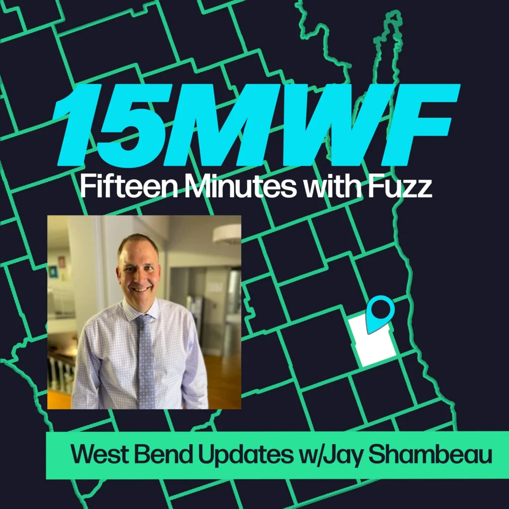 A graphic for “Fifteen Minutes with Fuzz” features a background map outline in green on a dark blue background. The title “15MWF” in large teal letters is at the top, with “Fifteen Minutes with Fuzz” in white letters underneath. Below the title, there is an inset photo of a man in a light-colored checkered shirt and tie, smiling at the camera in an indoor office setting. At the bottom, a green label reads “West Bend Updates w/Jay Shambeau.”