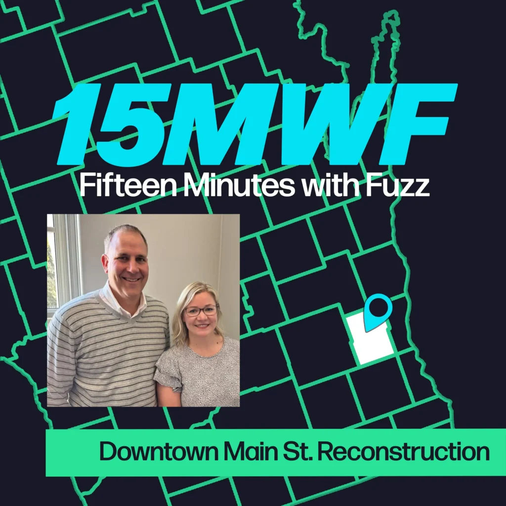 A graphic for “Fifteen Minutes with Fuzz” features a background map outline in green on a dark blue background. The title “15MWF” in large teal letters is at the top, with “Fifteen Minutes with Fuzz” in white letters underneath. Below the title, there is an inset photo of two people standing next to each other and smiling. One is a man wearing a striped sweater, and the other is a woman in a patterned shirt. At the bottom, a green label reads “Downtown Main St. Reconstruction.”