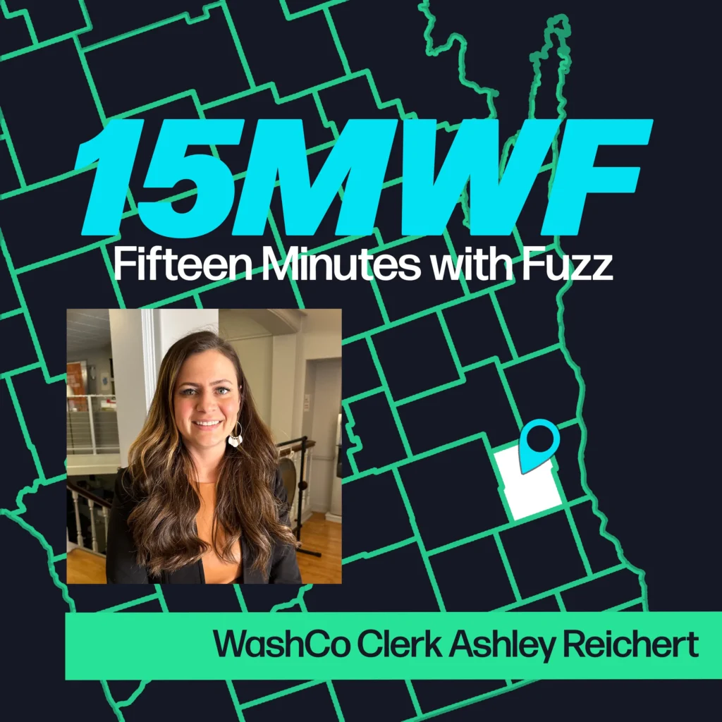 A graphic for “Fifteen Minutes with Fuzz” features a background map outline in green on a dark blue background. The title “15MWF” in large teal letters is at the top, with “Fifteen Minutes with Fuzz” in white letters underneath. Below the title, there is an inset photo of Ashley Reichert, wearing a black blazer and a brown top, with a white background. At the bottom, a green label reads “WashCo Clerk Ashley Reichert.”
