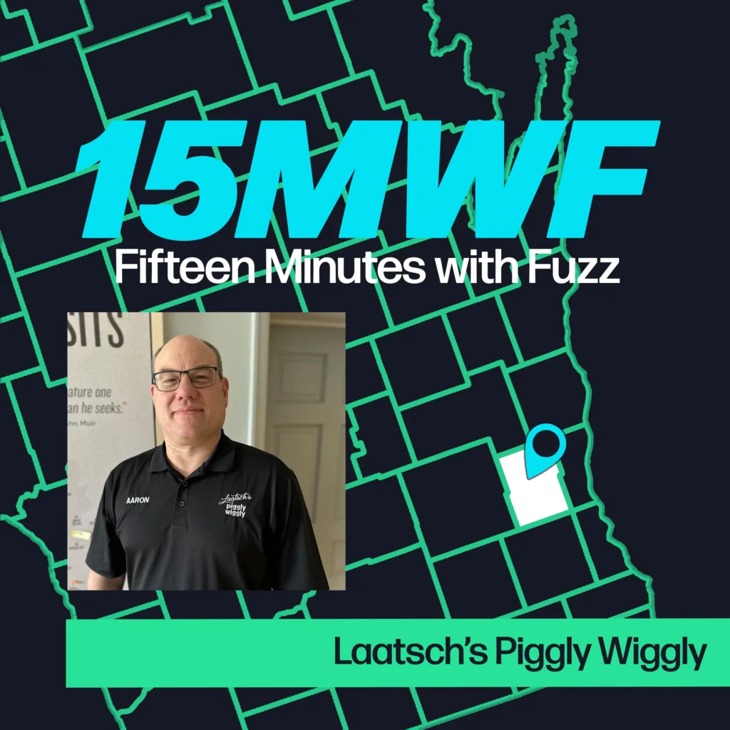 A graphic for “Fifteen Minutes with Fuzz” features a background map outline in green on a dark blue background. The title “15MWF” in large teal letters is at the top, with “Fifteen Minutes with Fuzz” in white letters underneath. Below the title, there is an inset photo of Aaron, wearing glasses and a black polo shirt with the name tag “Aaron” and “Laatsch’s Piggly Wiggly” logo on it. At the bottom, a green label reads “Laatsch’s Piggly Wiggly.”