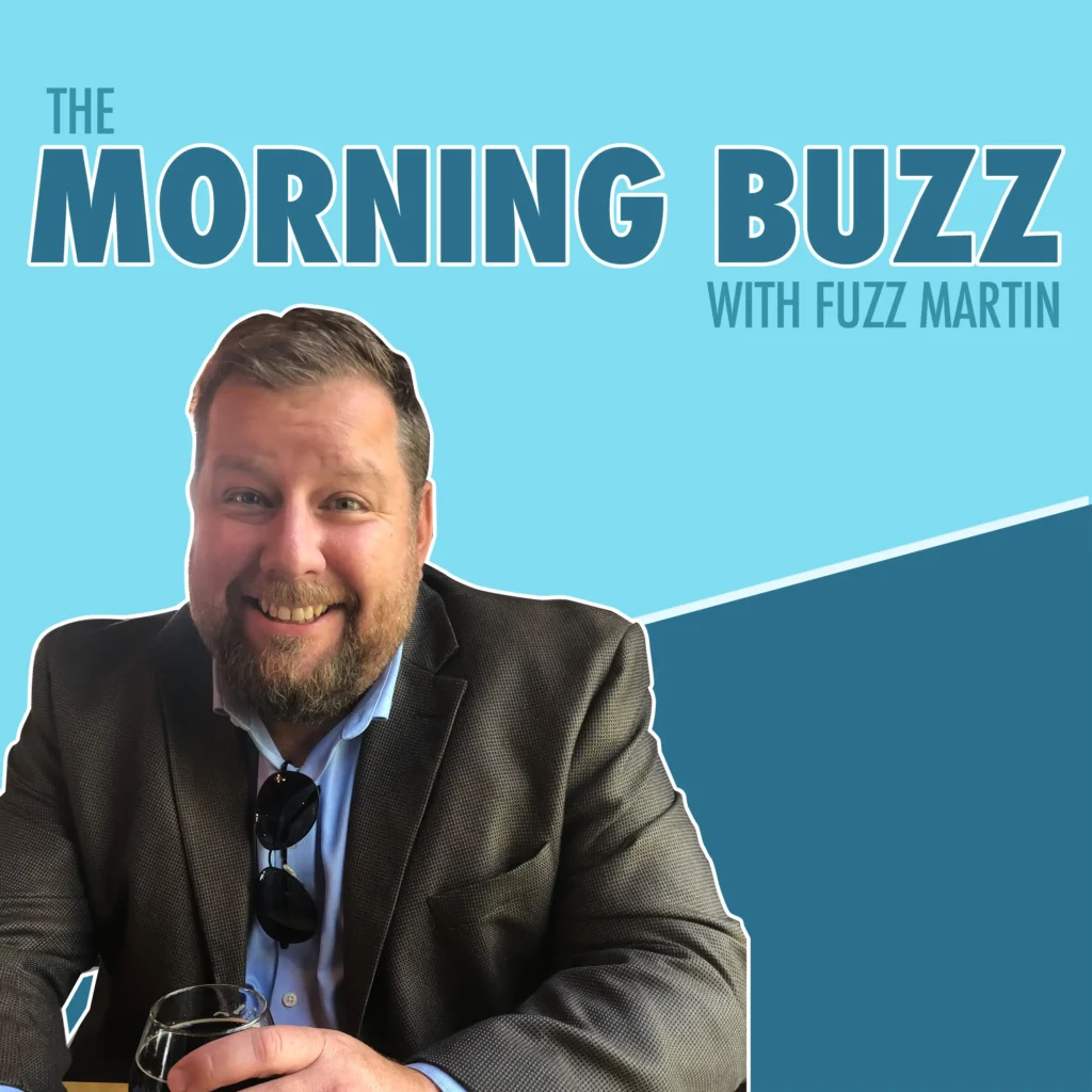 A promotional image for “The Morning Buzz with Fuzz Martin” features Fuzz Martin sitting at a table, smiling, and holding a drink. He is wearing a suit jacket over a light blue shirt with sunglasses hanging from the shirt’s collar. The background is a light blue gradient with a darker blue angular design cutting across the lower right corner. The title “THE MORNING BUZZ” is written in large, bold letters, with “WITH FUZZ MARTIN” in smaller text to the right.
