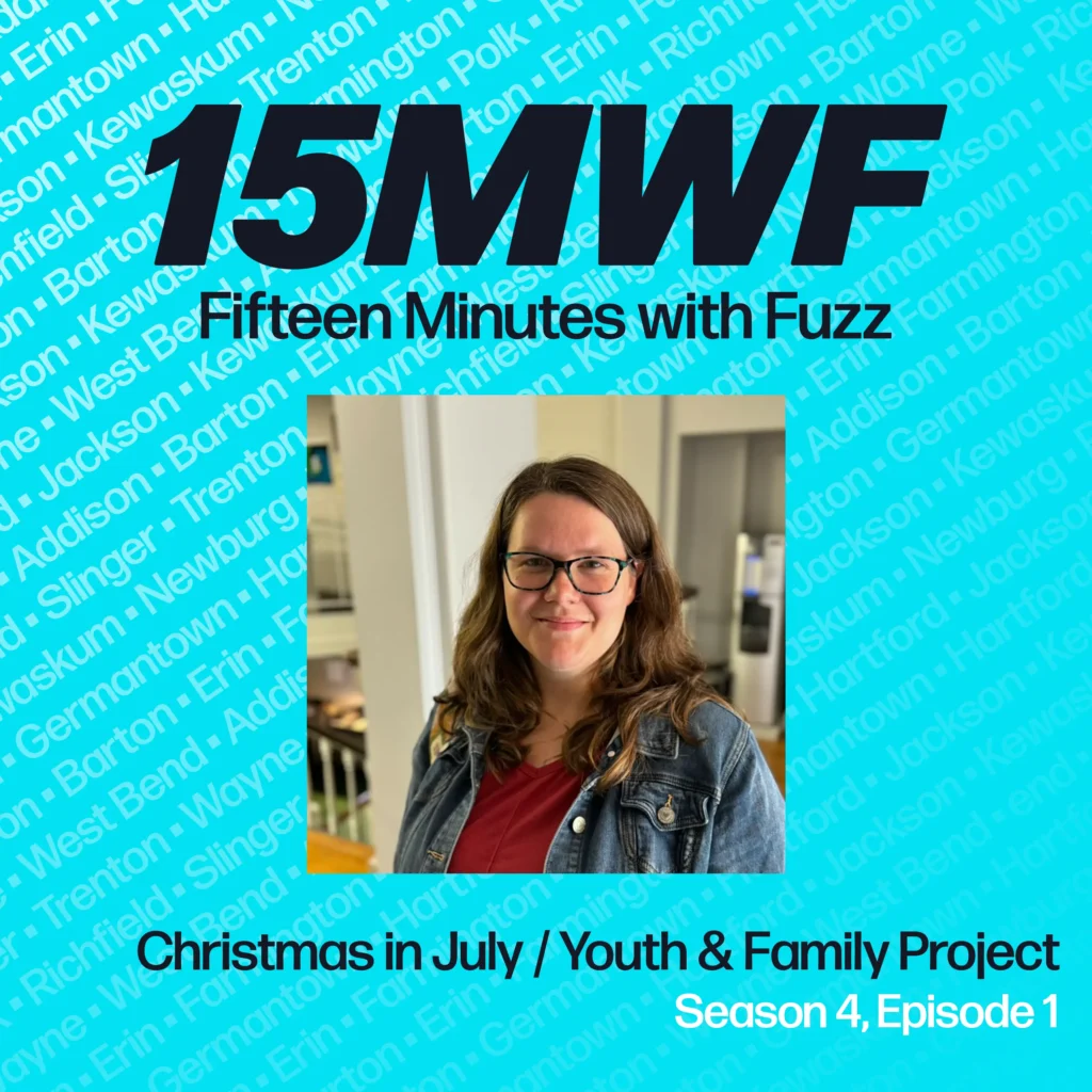 Promotional image for the podcast “Fifteen Minutes with Fuzz” featuring a headshot of a smiling woman with long brown hair and glasses, wearing a denim jacket and a red shirt. The background is a bright blue with various town names written in light blue text. The top of the image has the podcast logo “15MWF Fifteen Minutes with Fuzz,” and the bottom text reads “Christmas in July / Youth & Family Project, Season 4, Episode 1.”