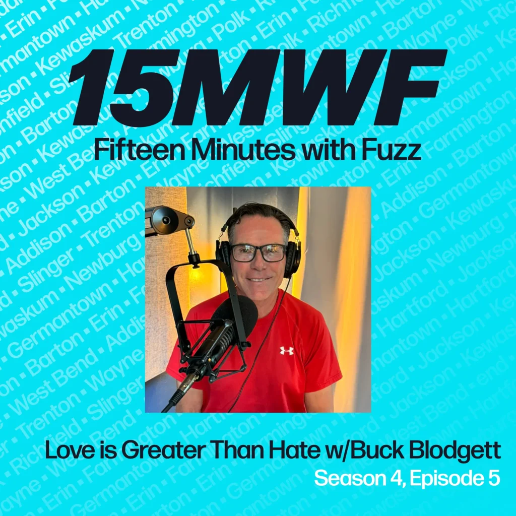 Promotional image for the ‘Fifteen Minutes with Fuzz’ podcast, featuring the text ‘15MWF’ in bold black letters and ‘Fifteen Minutes with Fuzz’ below it. Below the title, the episode title ‘Love is Greater Than Hate w/Buck Blodgett’ is displayed, along with ‘Season 4, Episode 5’. The right side of the image shows a photo of Buck Blodgett, wearing a red Under Armour shirt, sitting in front of a microphone with headphones on, smiling at the camera. The background features a light blue color with the names of various towns in a subtle pattern.